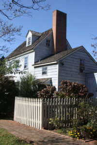 An exterior shot of a building with white siding, a grey roof, and a white fence, located inside the gardens.